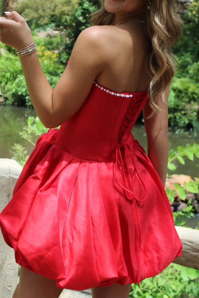 A woman modeling a light blue strapless homecoming mini dress with a structured bodice, flared skirt, and a large fabric rose detail on the side, standing against a white brick wall.