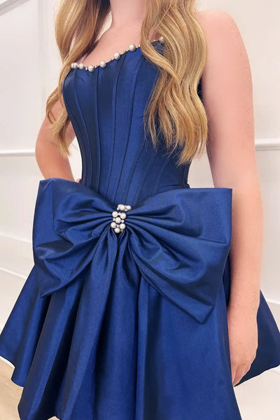 A woman models a short black strapless homecoming dress with a sweetheart neckline, pearl-like embellishments, a large bow at the waist, and a flared skirt, standing against a green hedge background.