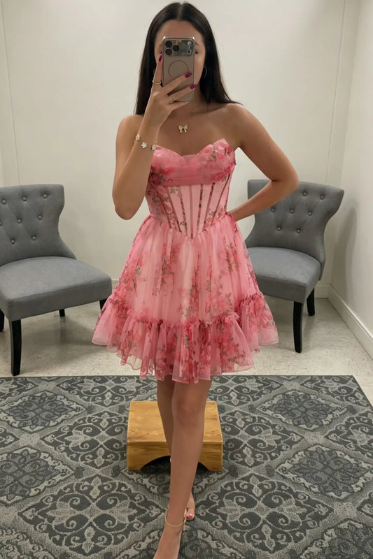 A woman takes a mirror selfie in a fitting room wearing a pink strapless tulle dress with a floral print and corset-style bodice. Two gray tufted chairs and a patterned rug are visible in the room.