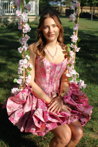 A woman wearing a strapless pink homecoming mini dress with a corset-style bodice and ruffled skirt, standing outdoors in front of a brick building.