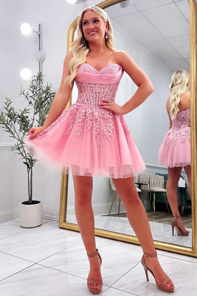A blonde woman models a short pink strapless tulle dress with a lace-embellished corset bodice in a bright indoor room with a large mirror.