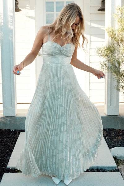 A woman stands on a front porch wearing a sleeveless, floor-length pleated gown in a very sage green color. The dress has thin straps, a fitted bodice, and a full gathered skirt.