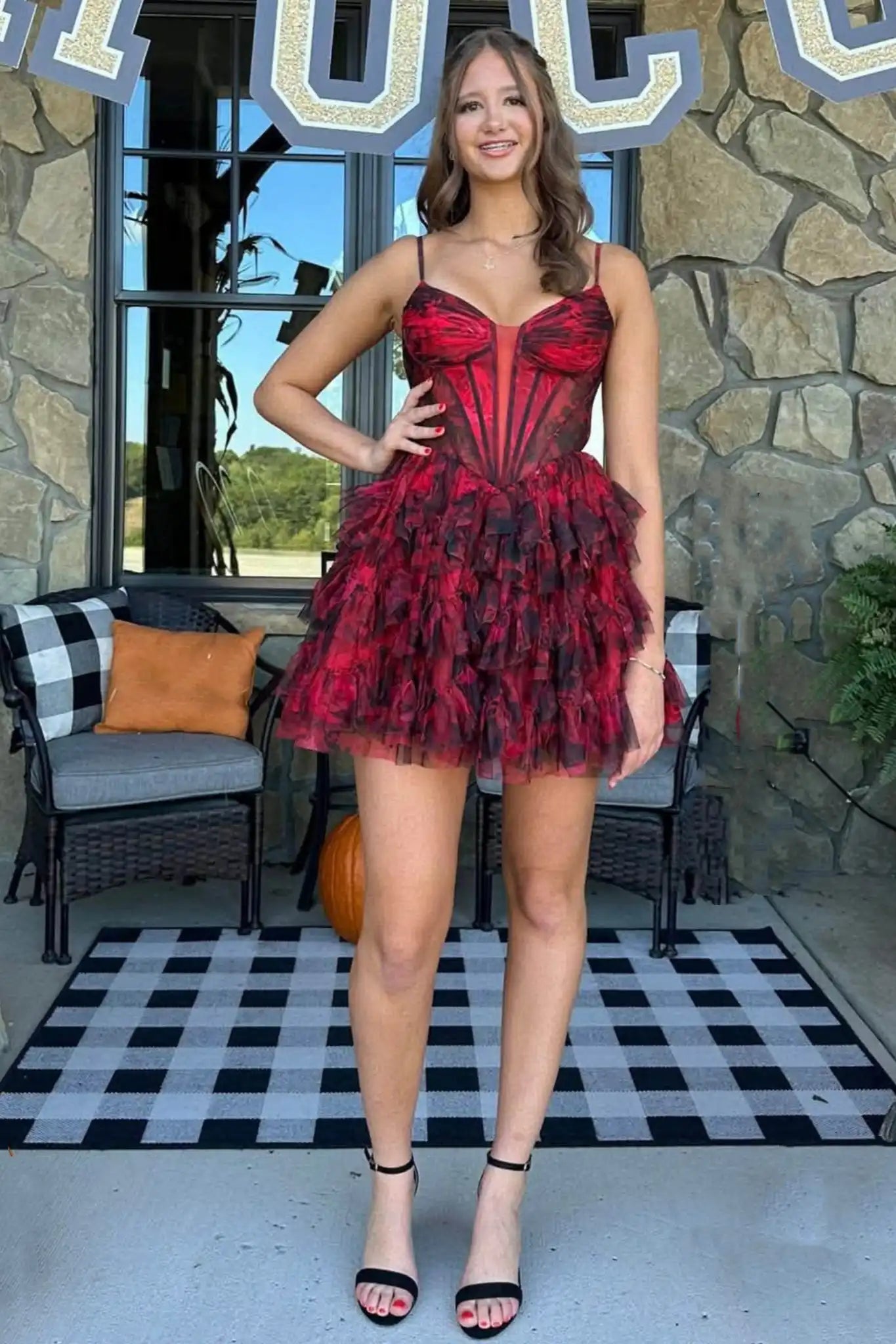 A woman modeling a short red and black ruffled tulle dress with a corset-style bodice, standing on a black-and-white checkered patio in front of a stone wall.