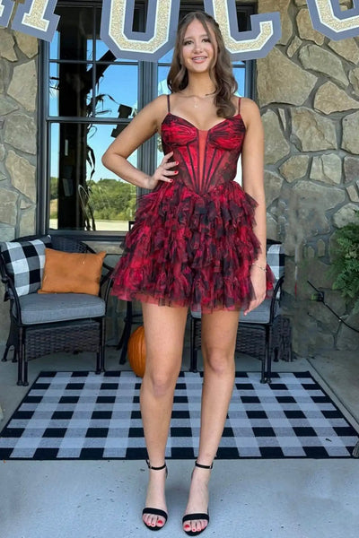 A woman modeling a short red and black ruffled tulle dress with a corset-style bodice, standing on a black-and-white checkered patio in front of a stone wall.