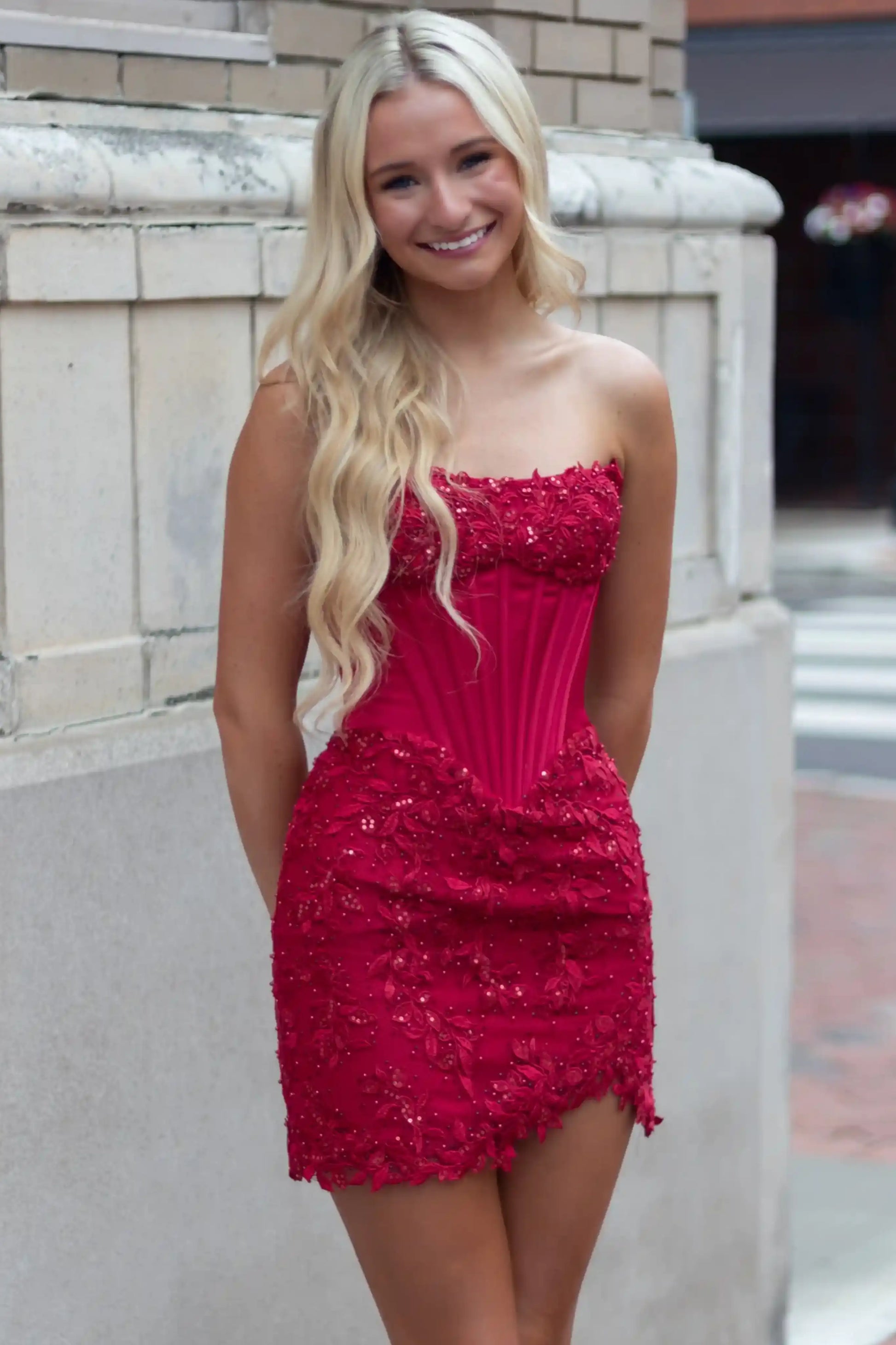 A smiling blonde woman wearing a bright red strapless mini dress with a corset-style bodice and beaded texture.