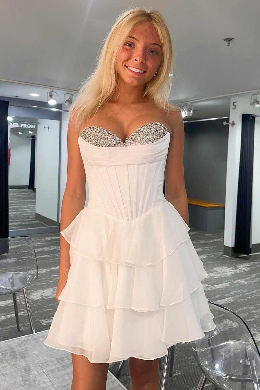 A blonde woman wearing a short white strapless dress with a rhinestone sweetheart neckline and a tiered ruffled skirt, standing in a fitting room.