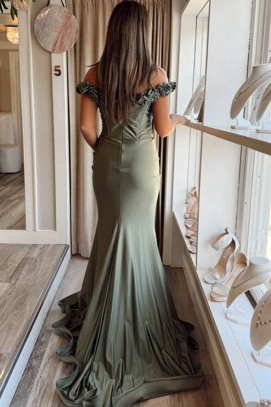 A woman viewed from behind wearing a floor-length off-the-shoulder mermaid gown in a muted sage green color, standing in a boutique with shoe displays and curtains in the background.