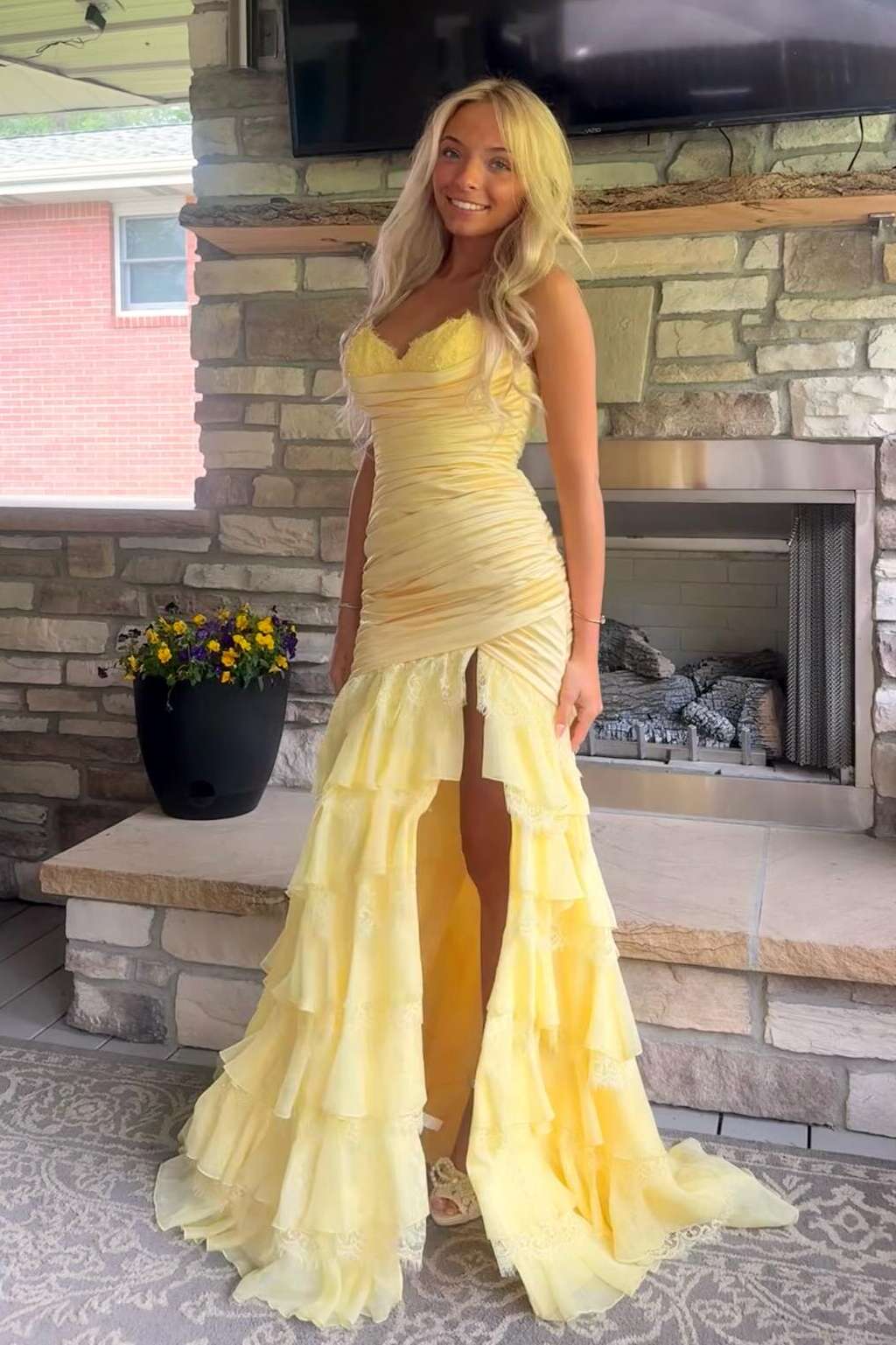 A woman wearing a yellow ruched mermaid-style prom gown with a high thigh slit and layered ruffle hem, standing in front of a stone fireplace.