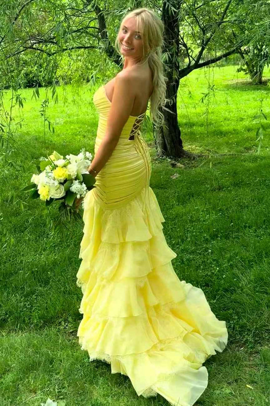 A woman stands outdoors on a grassy lawn in front of a tree, wearing a yellow strapless prom gown with a fitted ruched bodice and a layered ruffled skirt. 