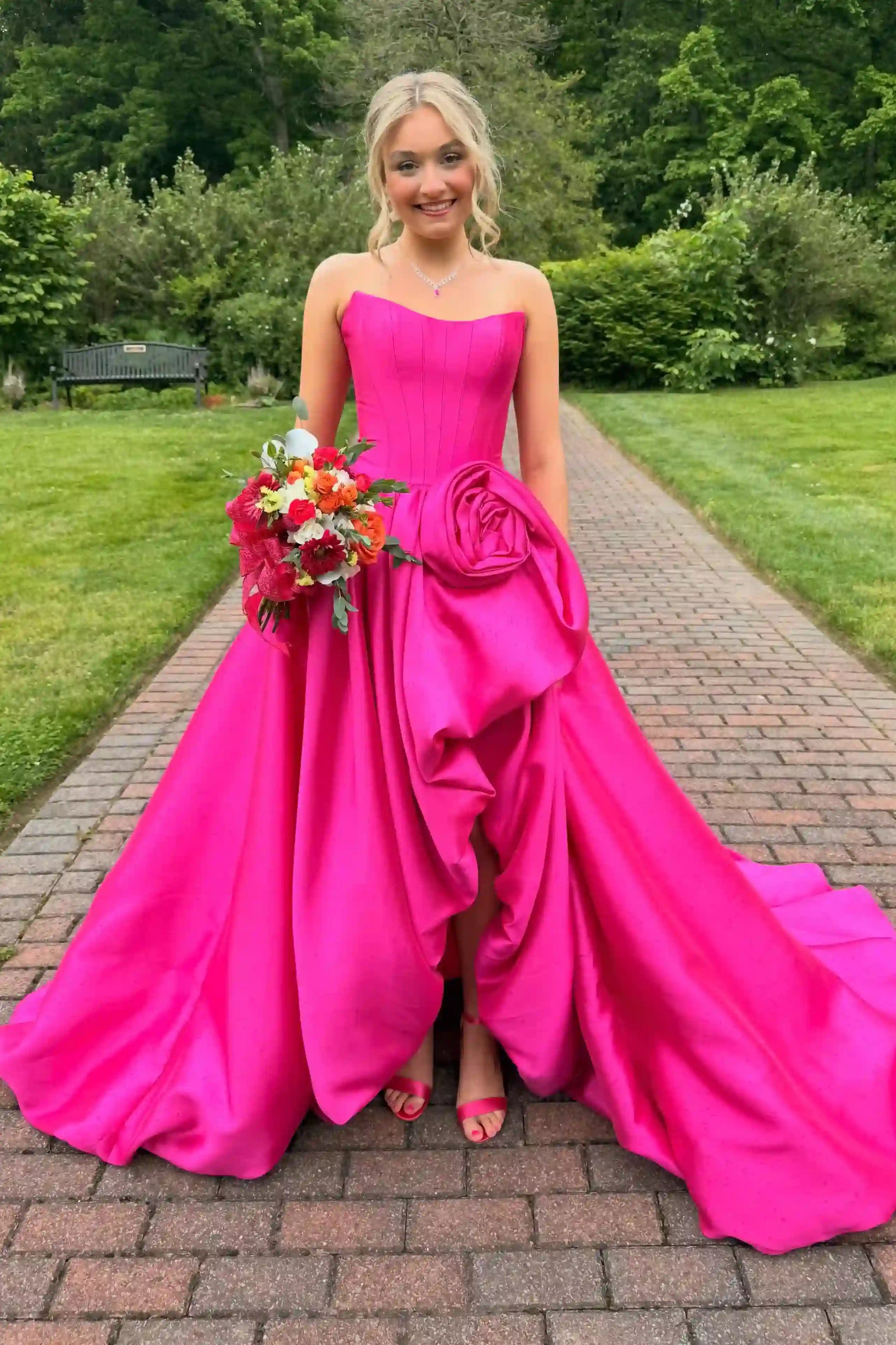 A woman stands on a brick path outdoors wearing a bright hot pink strapless gown with a large sculptural rose detail at the waist and a full flowing skirt. She is holding a bouquet of red and orange flowers, with green trees and grass in the background.
