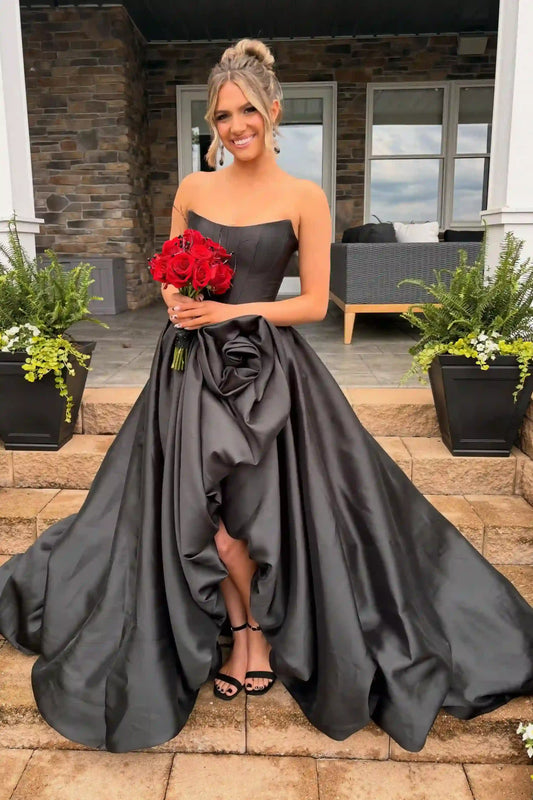 A woman stands on outdoor stone steps wearing a strapless black satin ball gown with a dramatic high-low hemline and a large gathered rosette at the waist. 