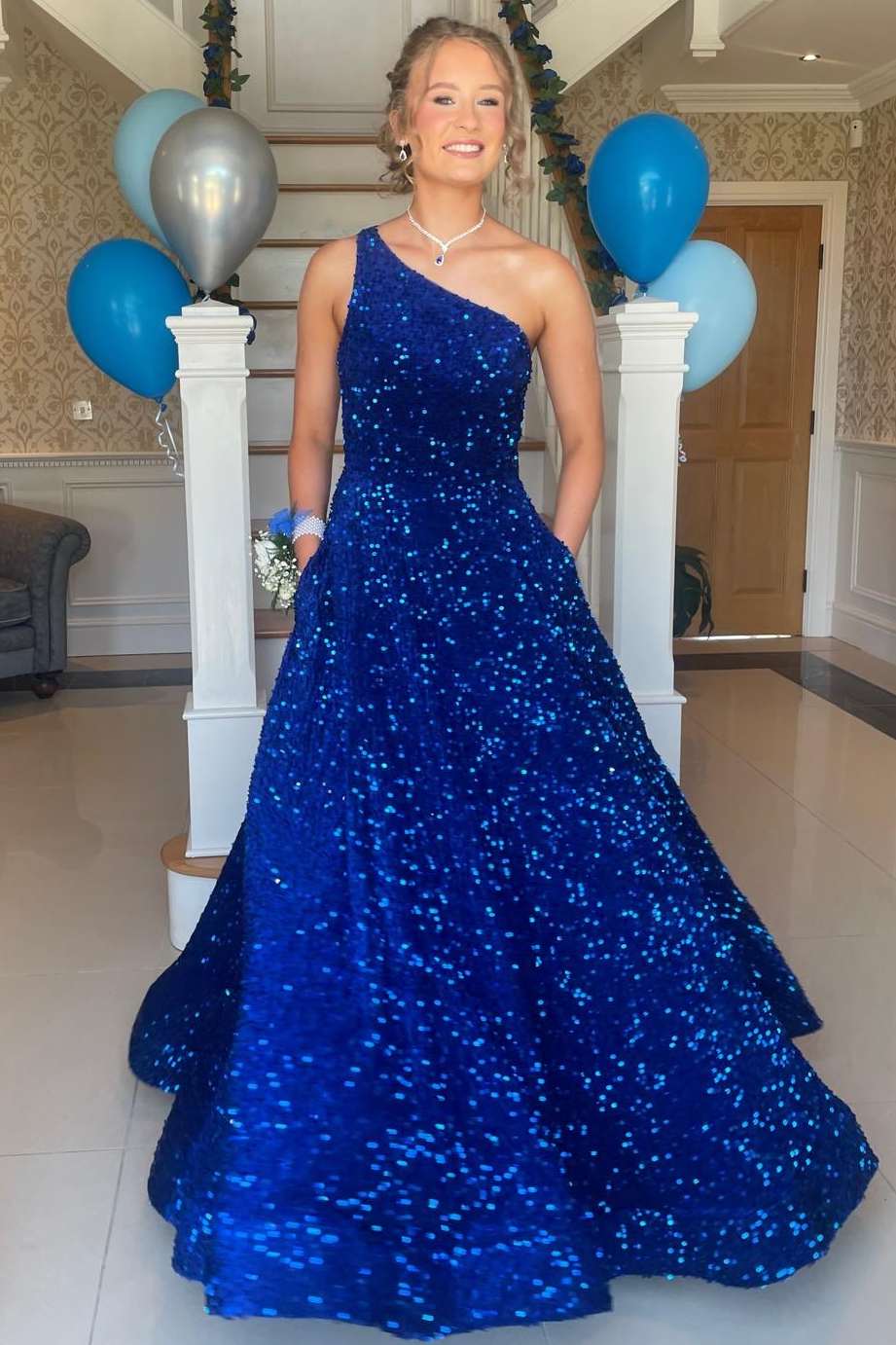 A woman wearing a royal blue sequined one-shoulder dress stands indoors in front of white pillars decorated with blue and silver balloons.