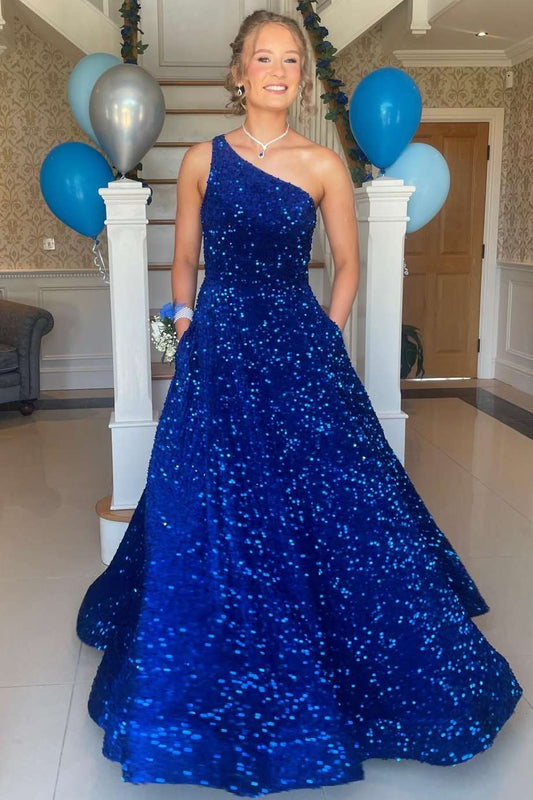 A woman wearing a royal blue sequined one-shoulder dress stands indoors in front of white pillars decorated with blue and silver balloons.