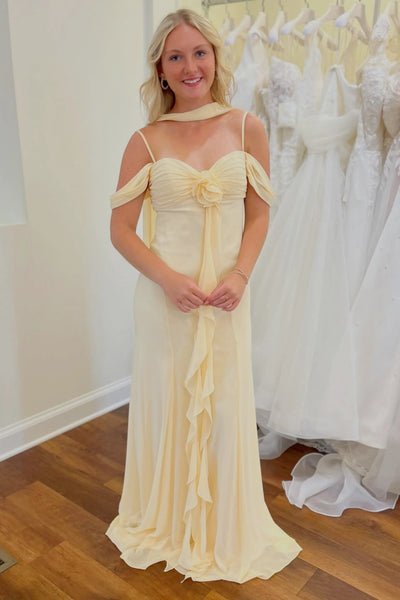 A woman modeling a yellow off-the-shoulder maxi bridesmaid dress with a ruffled front panel, standing in a bridal or formal wear shop with white dresses hanging in the background.