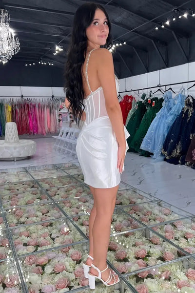 A woman modeling a short white satin mini dress with beaded spaghetti straps and a tie waist, standing in a bridal or formalwear showroom with dress racks and a flower-covered floor visible behind her.