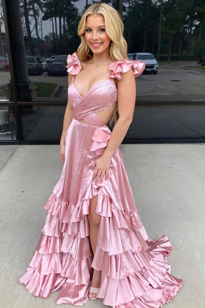 A blonde woman wearing a shiny light pink formal gown with ruffled shoulder details, a sweetheart neckline, and a tiered ruffle skirt, standing outdoors in front of a glass building.
