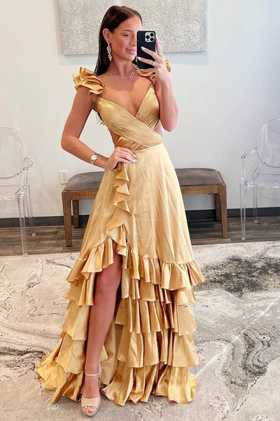 A blonde woman wearing a shiny light pink formal gown with ruffled shoulder details, a sweetheart neckline, and a tiered ruffle skirt, standing outdoors in front of a glass building.