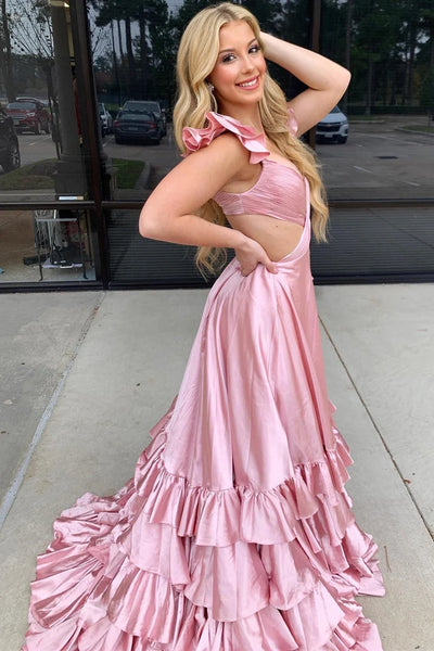 A blonde woman wearing a shiny light pink formal gown with ruffled shoulder details, a sweetheart neckline, and a tiered ruffle skirt, standing outdoors in front of a glass building.
