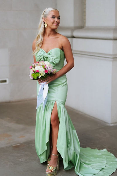 A woman wearing a strapless mint green mermaid-style gown with a sweetheart neckline, large bow detail at the bust, high-low skirt, thigh-high slit, and a short train, standing outdoors and holding a bouquet of pink and white flowers.