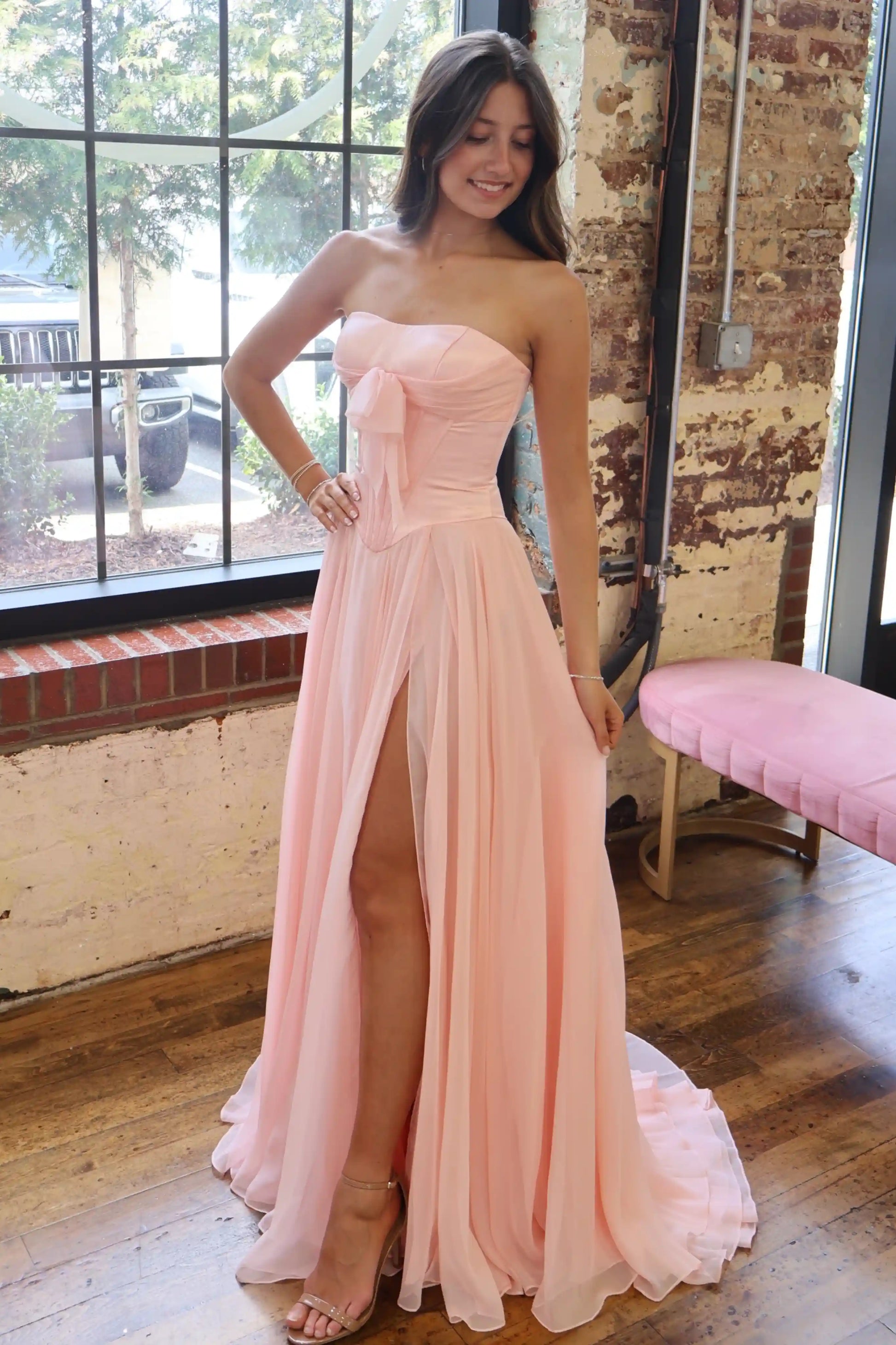 A woman models a strapless pink prom dress with a sweetheart neckline, a large bow detail at the waist, and a high thigh slit.