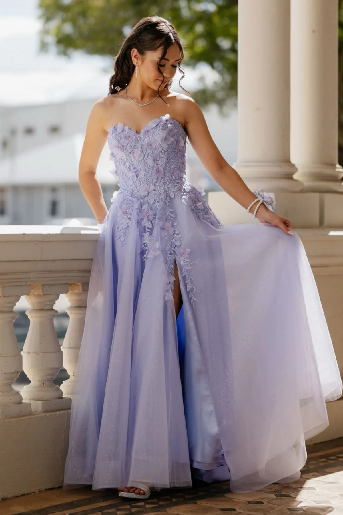 A woman models a strapless lavender tulle ball gown with floral applique on the bodice and skirt, featuring a thigh-high slit. She is standing on an outdoor porch with white columns and balustrades.