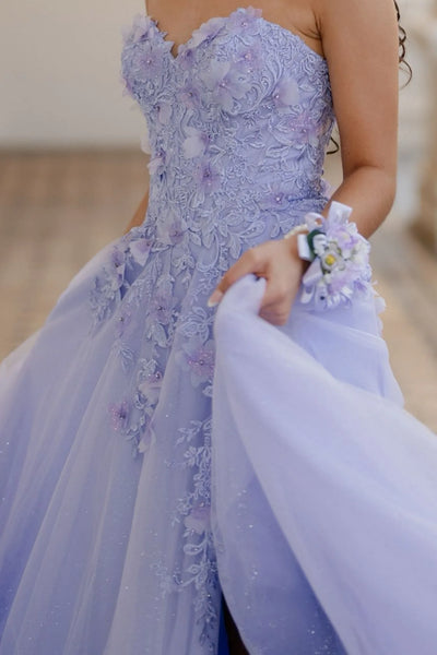 A woman models a strapless lavender tulle ball gown with floral applique on the bodice and skirt, featuring a thigh-high slit. She is standing on an outdoor porch with white columns and balustrades.