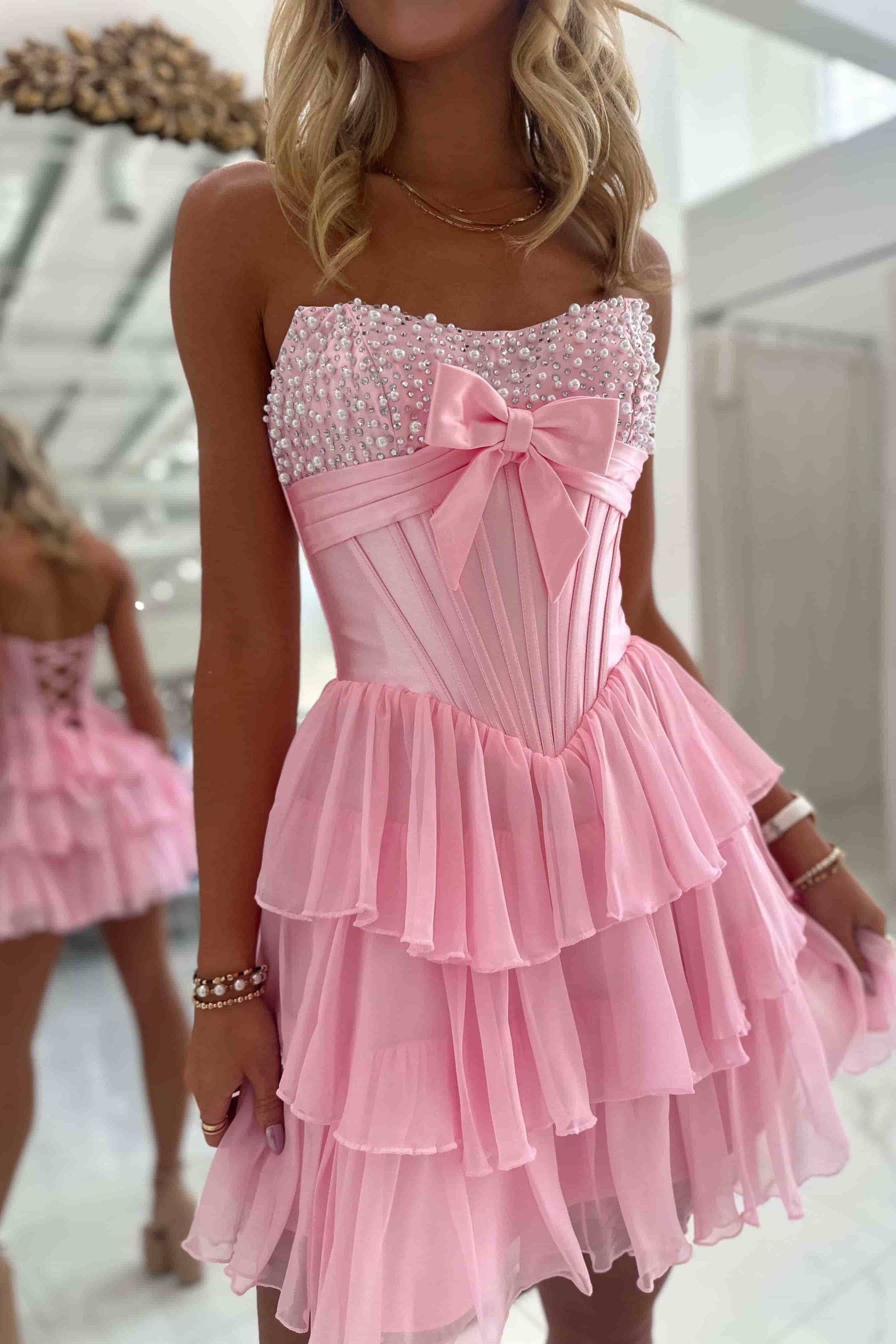 A pink strapless homecoming gown with a floral print and a large bow at the bust, displayed on a mannequin in a bright white room with paneled walls.