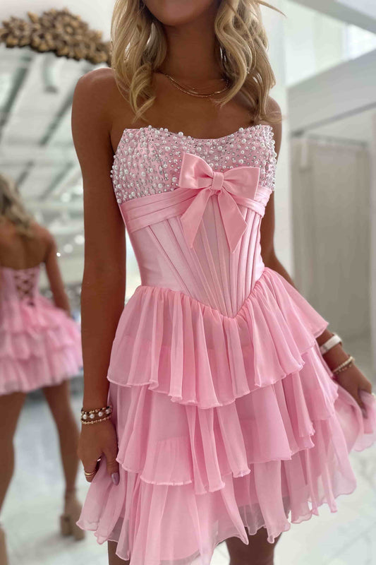 A pink strapless homecoming gown with a floral print and a large bow at the bust, displayed on a mannequin in a bright white room with paneled walls.