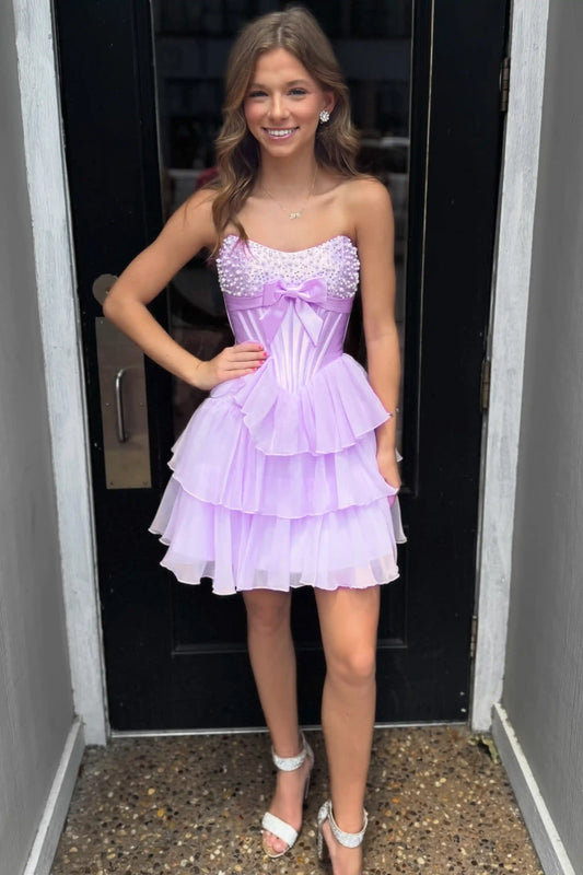 A young woman wearing a lavender purple strapless homecoming dress with a beaded sweetheart bodice, a large bow at the waist, and a tiered ruffled skirt, posing in front of a black door.