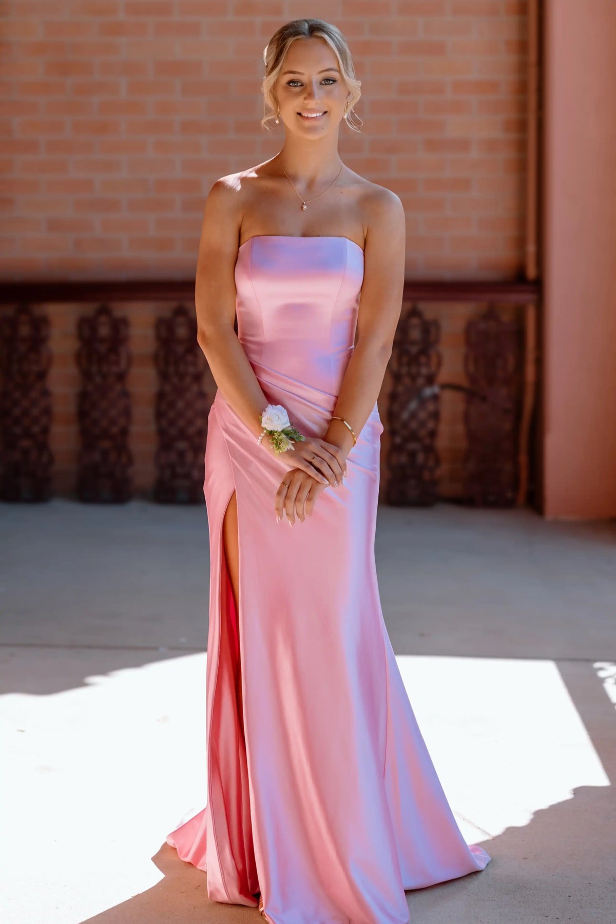 A woman wearing a strapless satin pink gown with a thigh-high slit, standing outdoors in front of a brick wall.