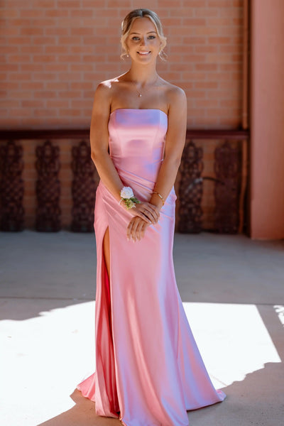 A woman wearing a strapless satin pink gown with a thigh-high slit, standing outdoors in front of a brick wall.