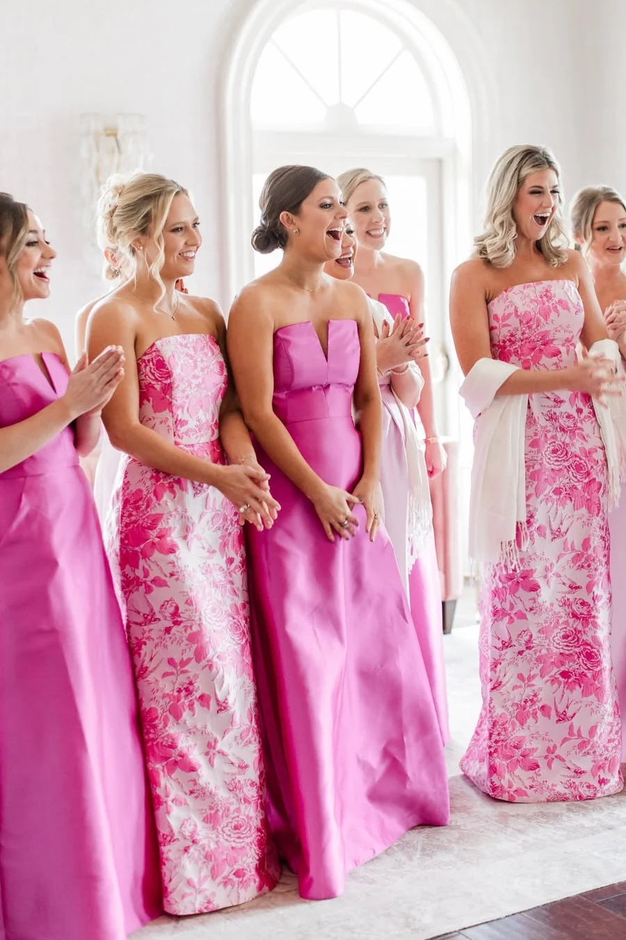 A group of bridesmaids wearing bright pink strapless maxi gowns with floral patterns, standing and laughing in a bright white indoor room with an arched window.
