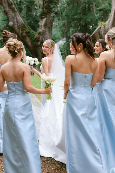 A group of bridesmaids wearing bright pink strapless maxi gowns with floral patterns, standing and laughing in a bright white indoor room with an arched window.