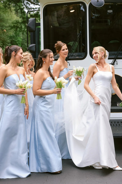 A group of bridesmaids wearing bright pink strapless maxi gowns with floral patterns, standing and laughing in a bright white indoor room with an arched window.