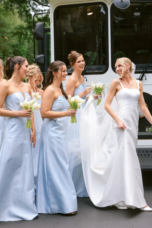 Bridesmaids in light blue strapless dresses and a bride in a white sleeveless gown walking beside a white bus outdoors, each holding white flower bouquets.