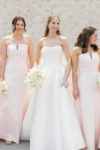 A group of bridesmaids wearing bright pink strapless maxi gowns with floral patterns, standing and laughing in a bright white indoor room with an arched window.
