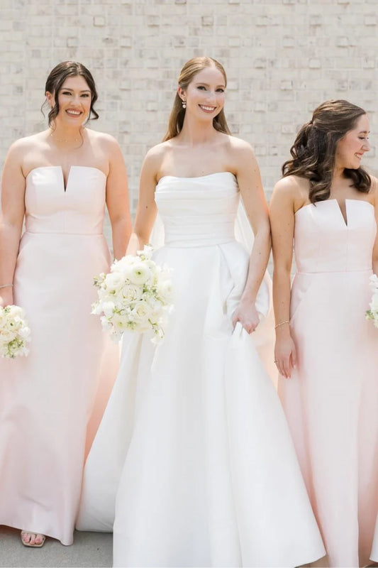 Three women standing together in formal wedding attire, with the central woman in a white strapless gown and the two women beside her wearing matching pale blush pink strapless bridesmaid dresses. The women are holding white flower bouquets against a light stone wall background.