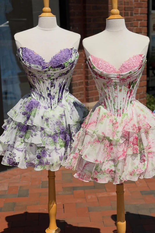 Two short corset dresses displayed on mannequins outdoors. The left dress is white with a lilac floral print and a purple lace bust.