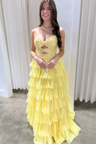 A woman wearing a bright yellow strapless gown with a sweetheart bodice, cutout details, and a tiered ruffled skirt, standing in front of cream-colored curtains.
