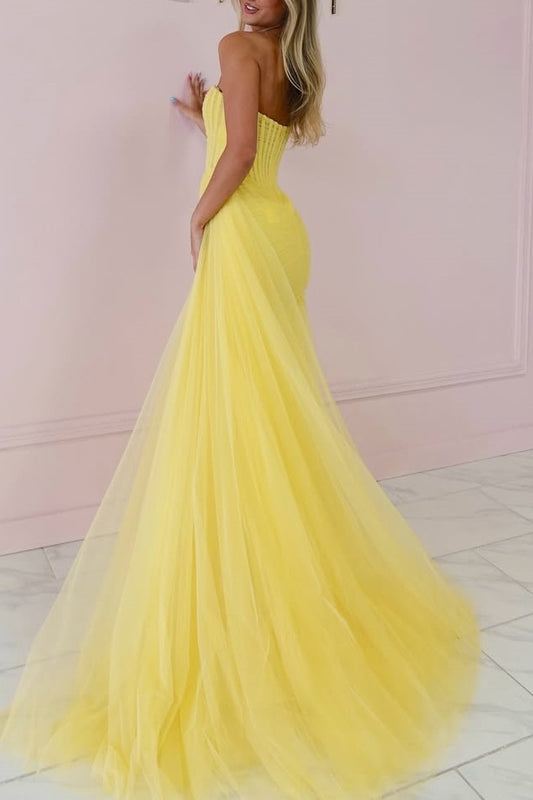 A woman modeling a strapless yellow tulle gown with a fitted bodice and full flowing skirt, photographed indoors against a pale pink wall and light tile floor.