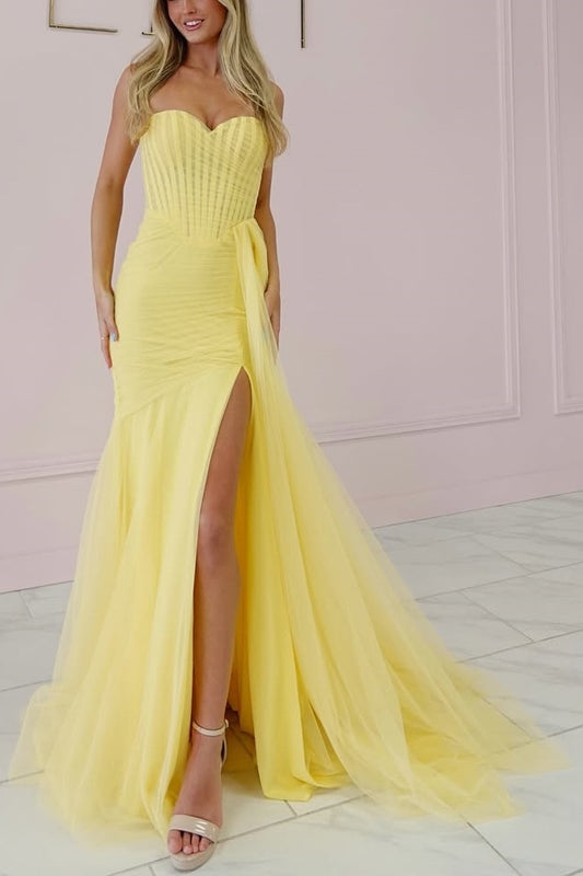 A woman models a strapless yellow mermaid gown with a sweetheart neckline, structured corset bodice, high thigh slit, and a flowing tulle train in a pale pink room.