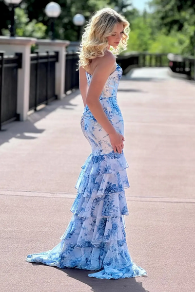 A woman stands outdoors on a pinkish-red walkway wearing a strapless formal gown with a blue and white floral print and a dramatic tiered ruffle skirt.