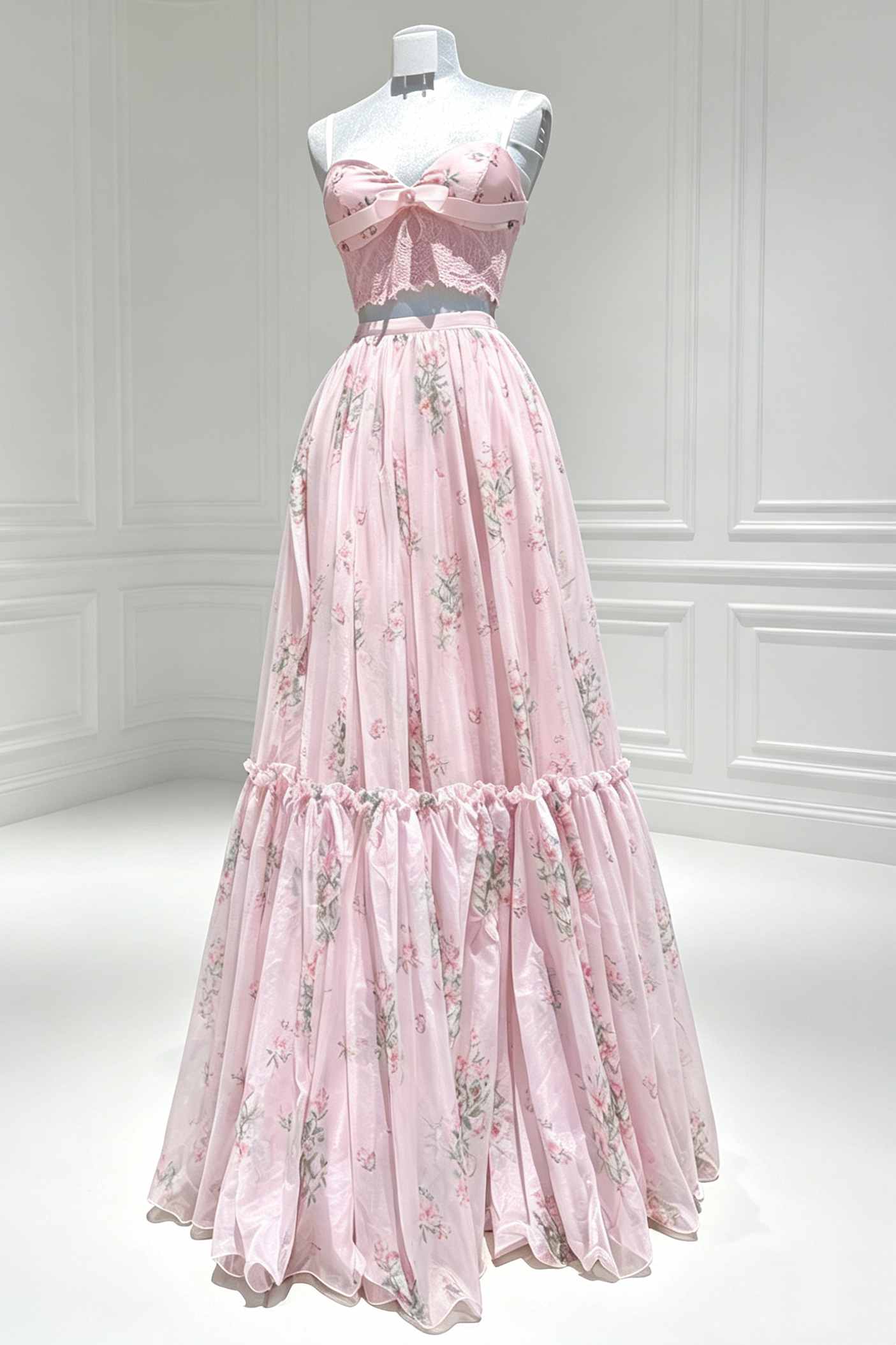A pink strapless formal gown with a floral print and a large bow at the bust, displayed on a mannequin in a bright white room with paneled walls.