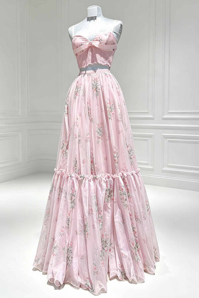 A pink strapless formal gown with a floral print and a large bow at the bust, displayed on a mannequin in a bright white room with paneled walls.