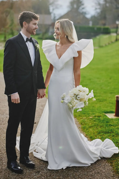 A bride in a white wedding dress with dramatic ruffled cap sleeves and a long train stands outdoors holding hands with a groom in a black tuxedo on a path beside a green lawn.