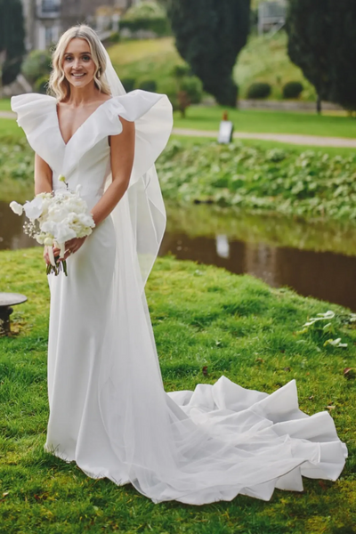 A bride in a white wedding dress with dramatic ruffled cap sleeves and a long train stands outdoors holding hands with a groom in a black tuxedo on a path beside a green lawn.
