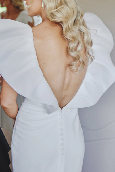 A bride in a white wedding dress with dramatic ruffled cap sleeves and a long train stands outdoors holding hands with a groom in a black tuxedo on a path beside a green lawn.