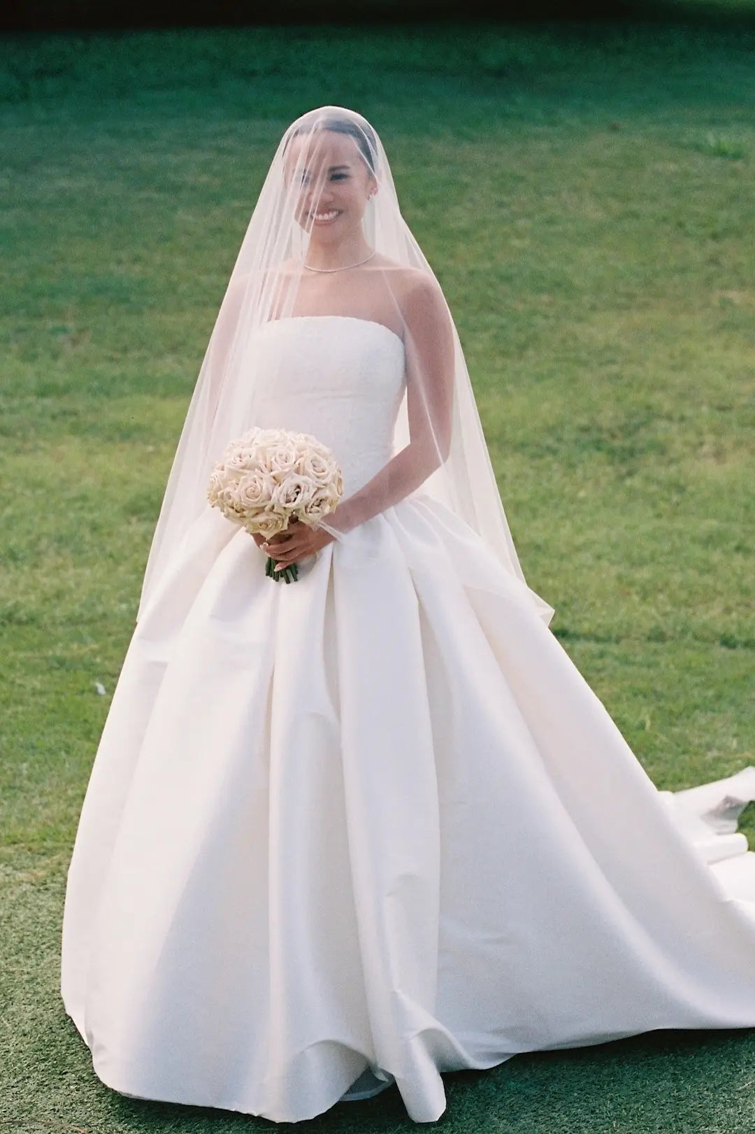 A woman wearing a strapless white wedding gown with a long sheer veil and holding a bouquet of cream flowers, standing outdoors on a green lawn.