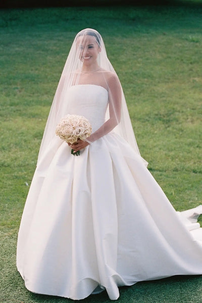 A woman wearing a strapless white wedding gown with a long sheer veil and holding a bouquet of cream flowers, standing outdoors on a green lawn.