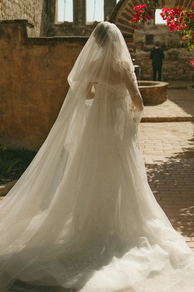 A bride in a fitted ivory lace wedding gown with a long sheer veil and a bouquet of white flowers, standing outdoors against a rustic stone wall.